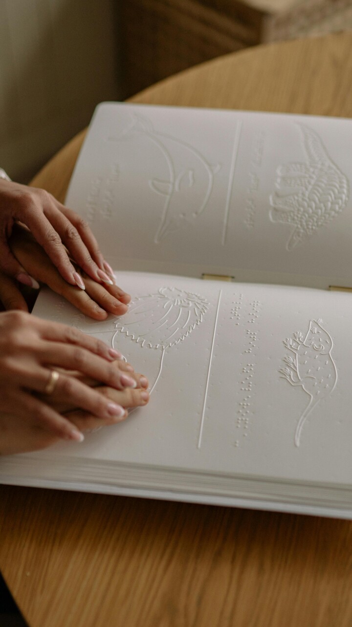 Ein Kind l&auml;sst seine H&auml;nde &uuml;ber ein Bilderbuch gleiten, dessen Motive gepr&auml;gt sind. Gef&uuml;hrt wird es dabei von den H&auml;nden einer erwachsenen Person.